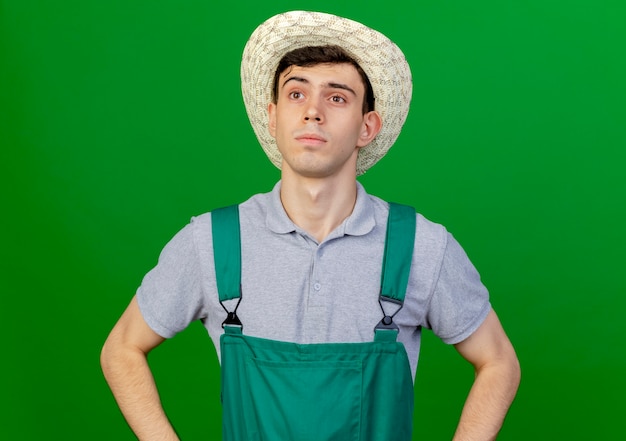 Foto gratuita jardinero de sexo masculino joven confiado con sombrero de jardinería pone las manos en la cintura mirando al lado aislado sobre fondo verde con espacio de copia