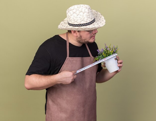 Jardinero de sexo masculino joven confiado con sombrero de jardinería que mide la flor en maceta con cinta métrica aislada en la pared verde oliva