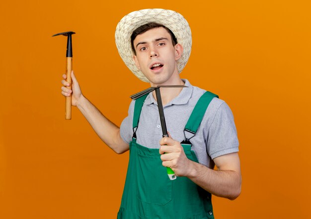 Jardinero de sexo masculino joven confiado con sombrero de jardinería tiene rastrillo y rastrillo de azada