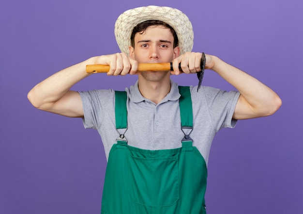Foto gratuita jardinero de sexo masculino joven confiado con sombrero de jardinería tiene rastrillo
