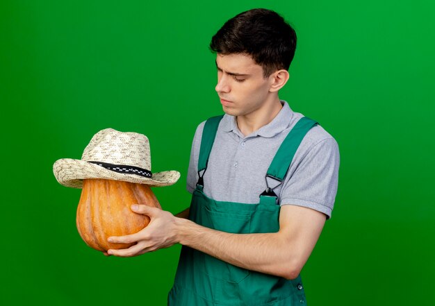 Jardinero de sexo masculino joven confiado sostiene y mira la calabaza con el sombrero de jardinería