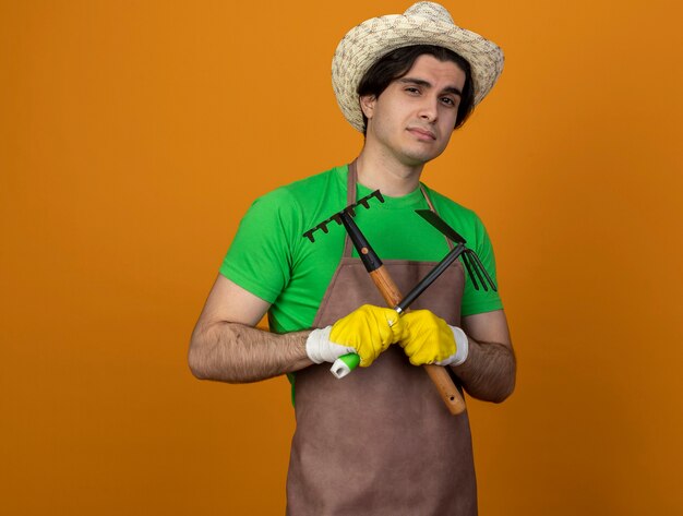 Foto gratuita jardinero de sexo masculino joven confiado en uniforme con sombrero de jardinería con guantes sosteniendo y cruzando rastrillo de azada con rastrillo aislado en naranja con espacio de copia