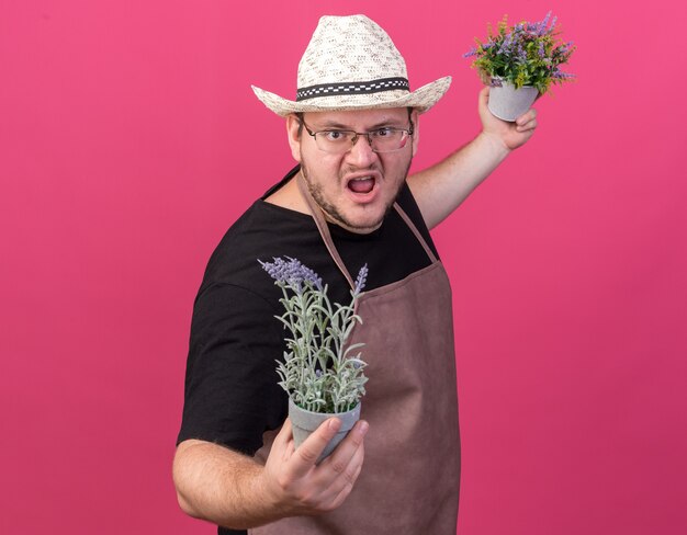 Jardinero de sexo masculino joven enojado con sombrero de jardinería sosteniendo flores en macetas aisladas en la pared rosa