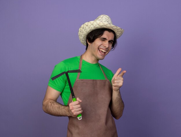 Jardinero de sexo masculino joven parpadeó en uniforme con sombrero de jardinería sosteniendo rastrillo de azada y mostrando gesto