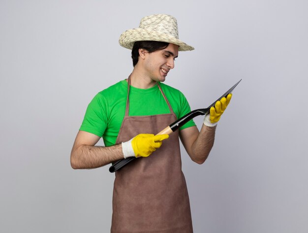 Jardinero de sexo masculino joven sonriente en uniforme que lleva el sombrero y los guantes de jardinería que sostienen y que miran la pala