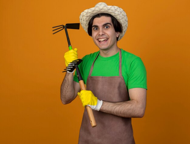 Jardinero de sexo masculino joven sonriente en uniforme que lleva el sombrero de jardinería con los guantes que sostienen el rastrillo de la azada con el rastrillo aislado en la pared naranja