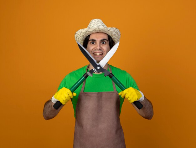 Jardinero de sexo masculino joven sonriente en uniforme que lleva el sombrero de jardinería con guantes que sujetan las tijeras