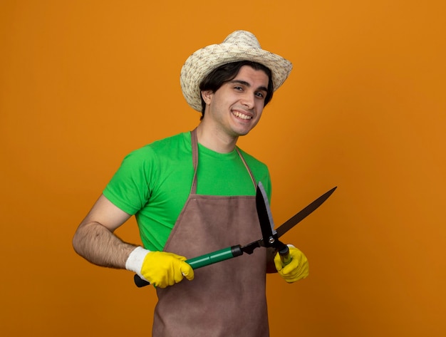 Jardinero de sexo masculino joven sonriente en uniforme que lleva el sombrero de jardinería con guantes que sujetan las tijeras