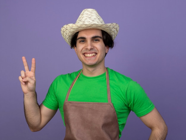 Jardinero de sexo masculino joven sonriente en uniforme que lleva el sombrero que cultiva un huerto que muestra el gesto de la paz aislado en púrpura