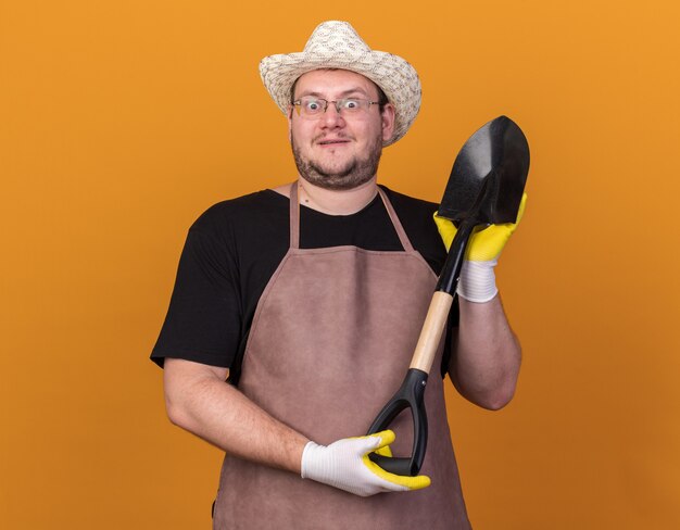 Jardinero de sexo masculino joven sorprendido que lleva el sombrero y los guantes de jardinería que sostienen la pala aislada en la pared anaranjada