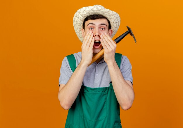 Jardinero de sexo masculino joven sorprendido que lleva el sombrero que cultiva un huerto pone las manos en la boca que sostiene el rastrillo