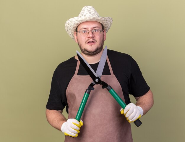 Jardinero de sexo masculino joven sorprendido con sombrero y guantes de jardinería sosteniendo tijeras aisladas en la pared verde oliva