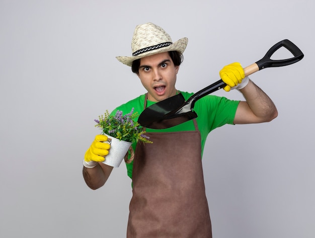 Jardinero de sexo masculino joven sorprendido en uniforme que lleva el sombrero de jardinería y los guantes que sostienen y puntos en flor en maceta con pala