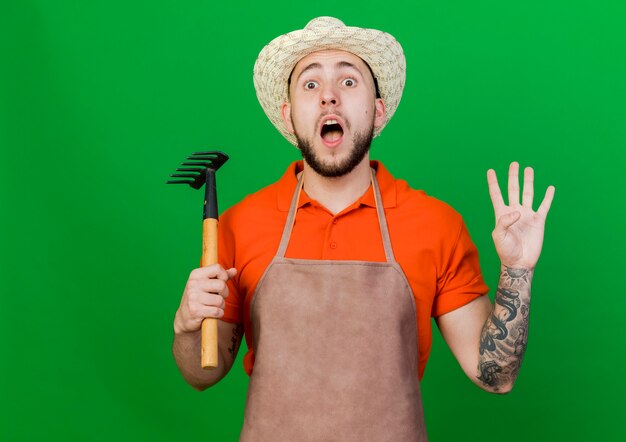 Foto gratuita jardinero sorprendido hombre vestido con sombrero de jardinería tiene rastrillo y gestos cuatro con los dedos