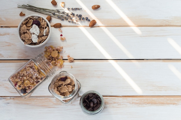 Foto gratuita jarra de granola derramada cerca de copos de maíz; frutas secas y chips de chocolate sobre tabla de madera.