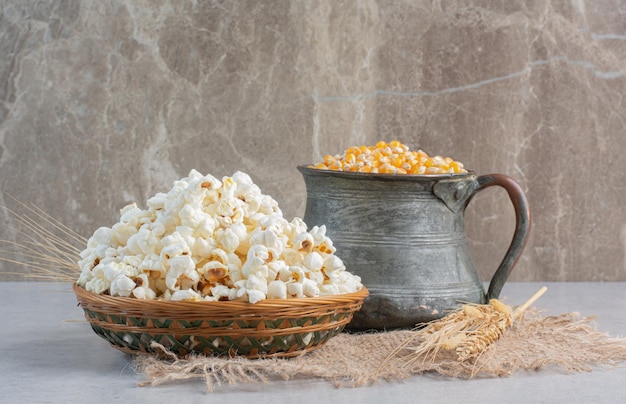 Una jarra de granos de maíz y un solo tallo de trigo junto a una canasta tejida de palomitas de maíz sobre un trozo de tela sobre la superficie de mármol