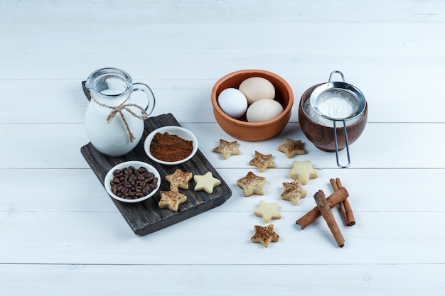 Jarra de leche, cuencos de granos de café y harina en una tabla de madera con galletas de estrella, canela, huevos, colador de harina vista de ángulo alto sobre un fondo de tablero de madera blanca