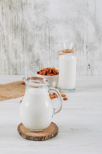 Jarra de leche con tazón de almendras y botella de leche vista de ángulo alto sobre un fondo blanco de madera y un saco