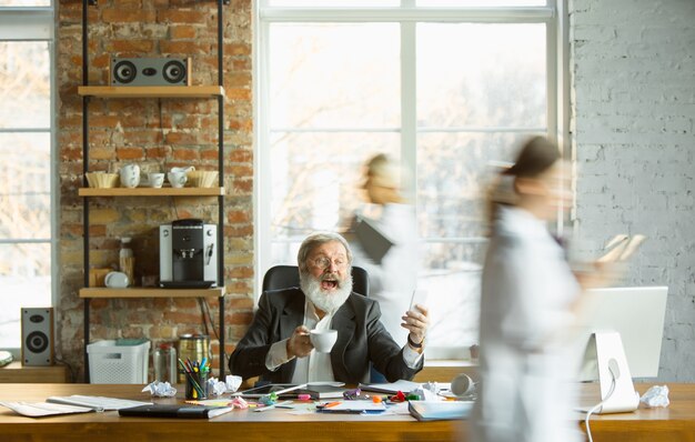 Jefe cansado descansando en su lugar de trabajo mientras la gente ocupada moviéndose cerca borrosa. Oficinista, gerente trabajando, tomando café y dando instrucciones a sus colegas. Negocio, trabajo, concepto de carga de trabajo.