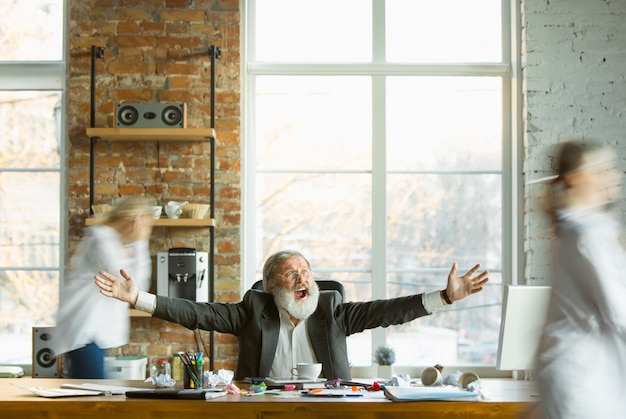 Jefe cansado descansando en su lugar de trabajo mientras la gente ocupada moviéndose cerca borrosa. Oficinista, gerente trabajando, tomando café y dando instrucciones a sus colegas. Negocio, trabajo, concepto de carga de trabajo.