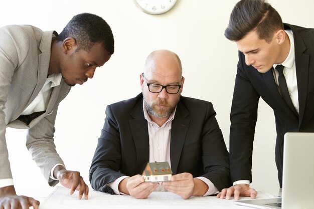 Jefe caucásico en vasos sosteniendo la casa modelo a escala del futuro inmobiliario mientras dos jóvenes arquitectos le presentan el proyecto de construcción.