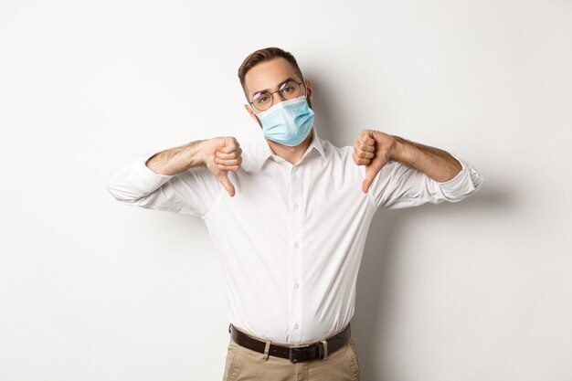 Jefe disgustado con mascarilla y mostrando los pulgares hacia abajo, signo de desaprobación y disgusto, fondo blanco.