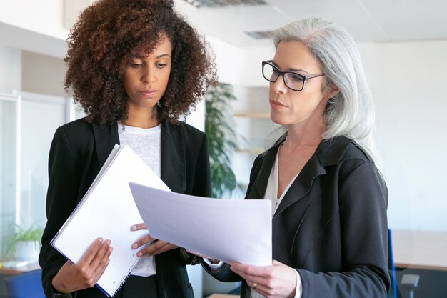 Jefe femenino confiado enfocado en vidrios que lee el informe. Empresaria joven acertada atractiva afroamericana que sostiene la documentación para el gerente. Concepto de trabajo en equipo, negocios y gestión