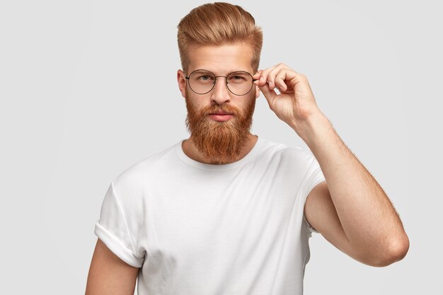 Jefe de hombre de moda serio con espesa barba pelirroja y peinado, toca el borde de las gafas