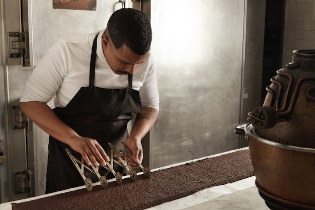 Foto gratuita el jefe de hombre negro de vista lateral usa un separador vintage profesional para dividir el pastel de chocolate en porciones iguales antes del empaque, proceso de cocción artesanal en confitería