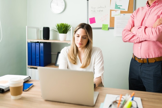 Jefe masculino molesto de pie junto a una compañera de trabajo y revisando su trabajo. Hermosa mujer caucásica trabajando y escribiendo en una computadora portátil en el escritorio de la oficina