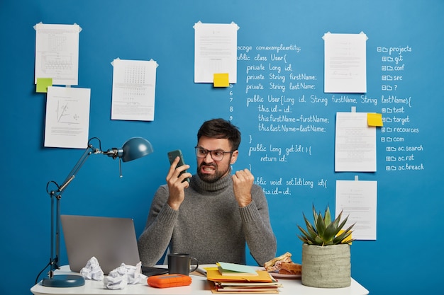 El jefe masculino molesto trabaja en casa en un lugar de trabajo acogedor, organiza el proceso de trabajo, aprieta el puño y los dientes con irritación