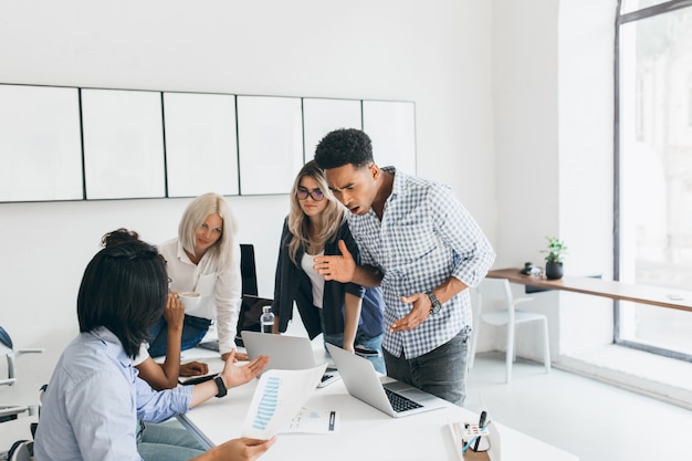 Foto gratuita jefe negro enojado de pie junto a la mesa y agitando las manos. retrato interior del gerente infeliz mirando la pantalla del portátil y diciéndole algo al programador asiático con camisa azul.