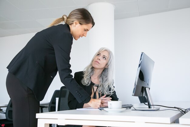 Jefe de proyecto que coloca la firma en el informe de los empleados. Compañeros de trabajo femeninos sentados y de pie en el lugar de trabajo con monitor y taza de café. Concepto de comunicación empresarial