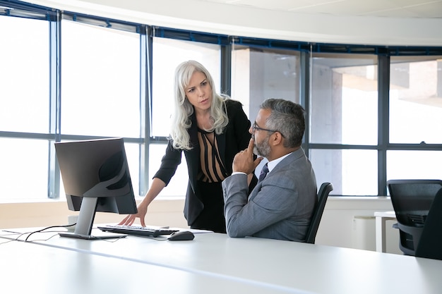 Jefe serio y gerente discutiendo el informe, hablando mientras está sentado y de pie en el lugar de trabajo con ventana panorámica. Concepto de comunicación empresarial