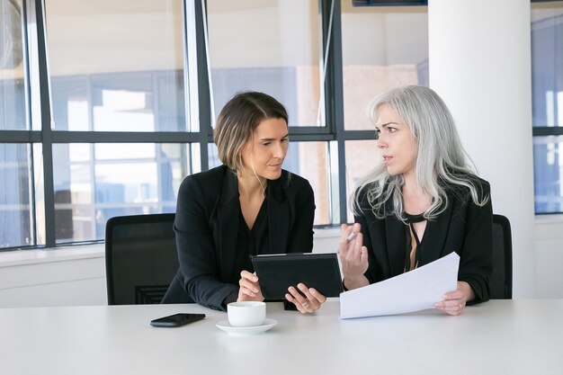 Jefes de proyecto analizando informes. Dos compañeras de trabajo sentadas juntas, mirando documentos, usando tableta y hablando. Vista frontal. Concepto de comunicación