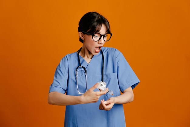 Jeringa de sujeción asustada joven doctora vistiendo uniforme estetoscopio fith aislado sobre fondo naranja