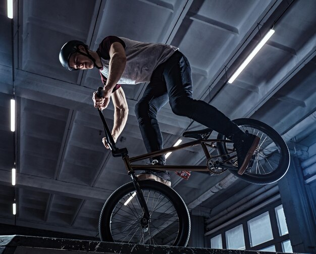 Jinete profesional de BMX en casco protector realizando trucos en skatepark en interiores