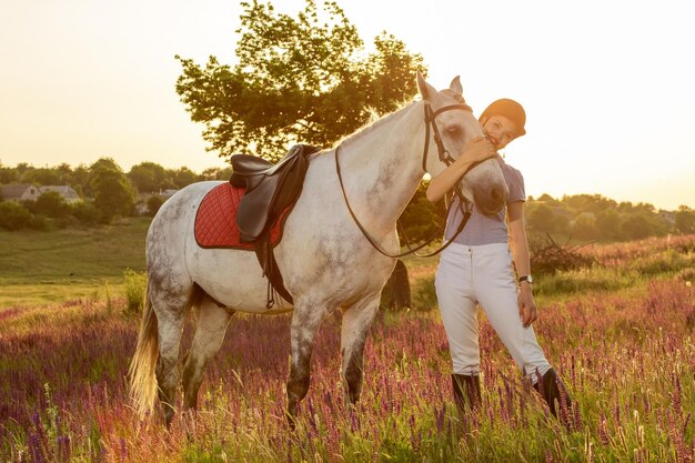 Jockey joven acariciando y abrazando al caballo blanco en el atardecer. llamarada del sol, destello solar