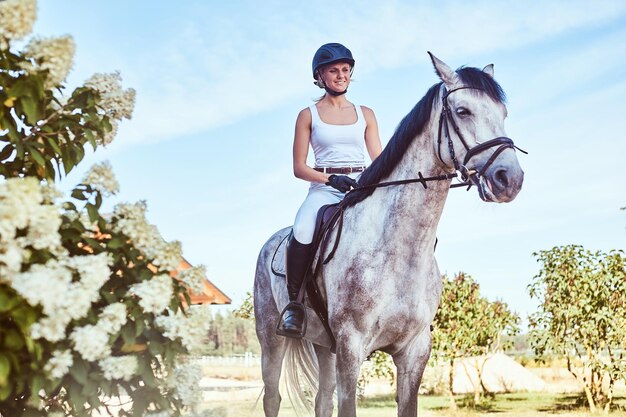 Una jockey sonriente sobre un caballo gris moteado caminando por el jardín florido.