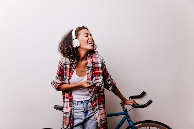 Foto gratuita jocund modelo femenino en auriculares de pie junto a la bicicleta. filmación en interiores de hermosa mujer rizada en camisa a cuadros roja.