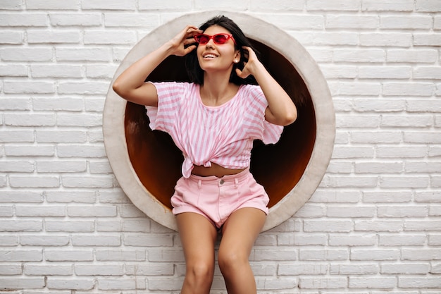 Foto gratuita jocund mujer bronceada riendo sobre fondo urbano. mujer emocionada sentada en la pared de ladrillo blanco.