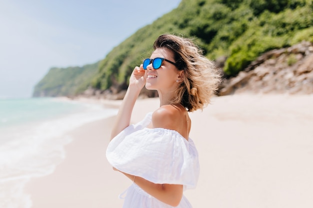 Foto gratuita jocund mujer joven en vestido y gafas de sol mirando al océano. bonita modelo femenina con piel bronceada pasando el fin de semana en el resort.