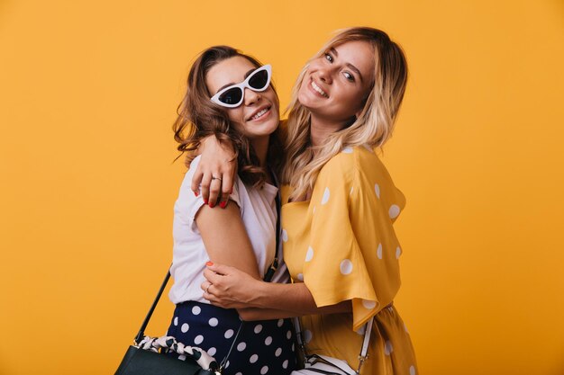 Jocund mujer rubia con vestido amarillo abrazando a su mejor amiga Inspirada dama de cabello oscuro con gafas de sol relajándose con su hermana