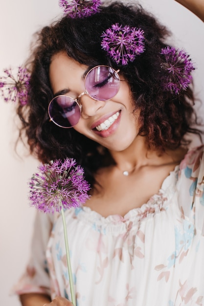 Jocund niña africana posando con alliums y riendo. Filmación en interiores de elegante mujer negra con cabello ondulado sonriendo.