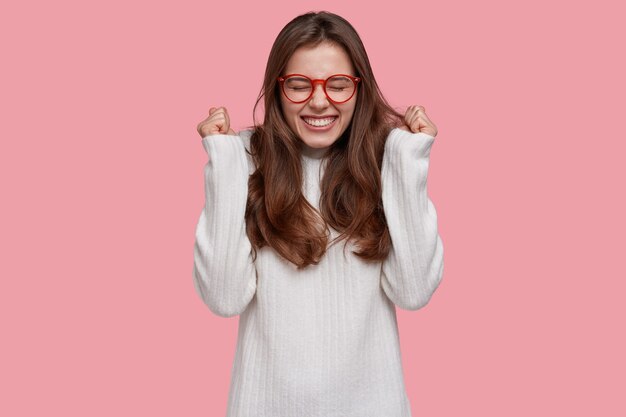 Foto gratuita joful triunfante joven aprieta los puños en gesto de victoria, sonríe de satisfacción, se regocija por la victoria, tiene el pelo largo y oscuro