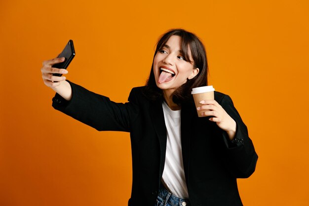 Jokin sosteniendo una taza de café, tome una selfie joven y hermosa mujer con chaqueta negra aislada en el fondo naranja