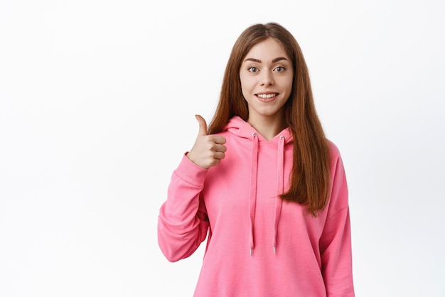 Una joven de 20 años con una sudadera con capucha informal que muestra los pulgares hacia arriba y sonríe le da su aprobación y está de acuerdo en que es bueno que elogie su elección de pie sobre un fondo blanco