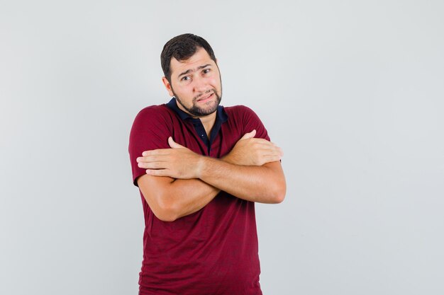 Joven abrazándose a sí mismo en camiseta roja y mirando frío, vista frontal.