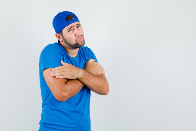 Joven abrazándose a sí mismo o sintiendo frío en camiseta azul y gorra y luciendo humilde