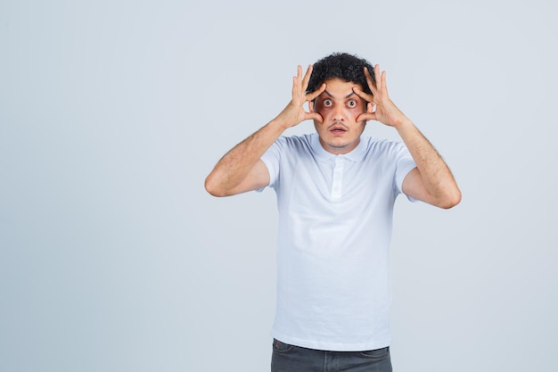 Joven abriendo los ojos con las manos en camiseta blanca, pantalones y mirando enfocado, vista frontal.